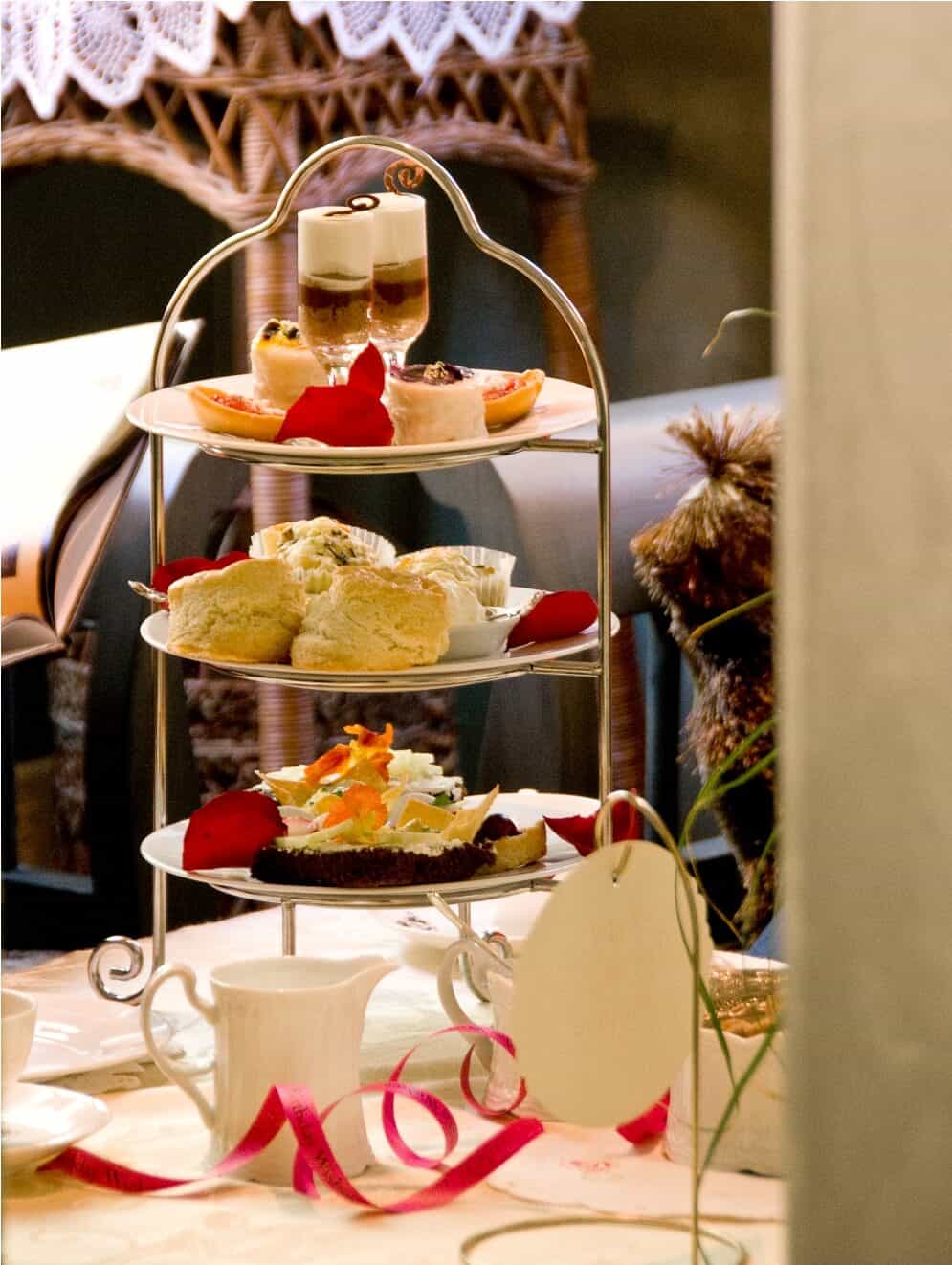 A three-tiered tray holds assorted pastries, scones, and desserts, decorated with rose petals, on a table set for tea with white cups and a teapot. A pink ribbon and blank tag are also on the table.