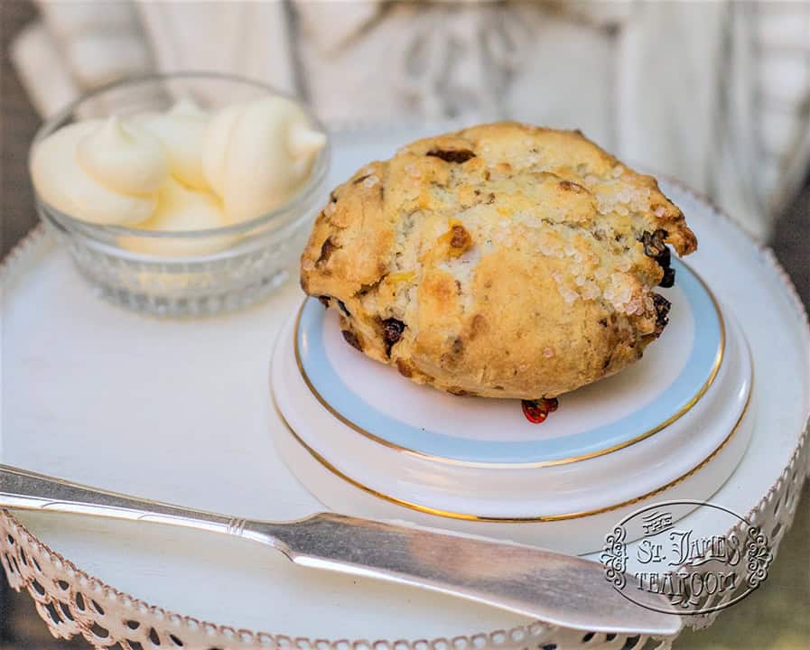 A golden scone dotted with raisins sits on a small plate beside a dish of clotted cream, with a butter knife resting on a white tray.