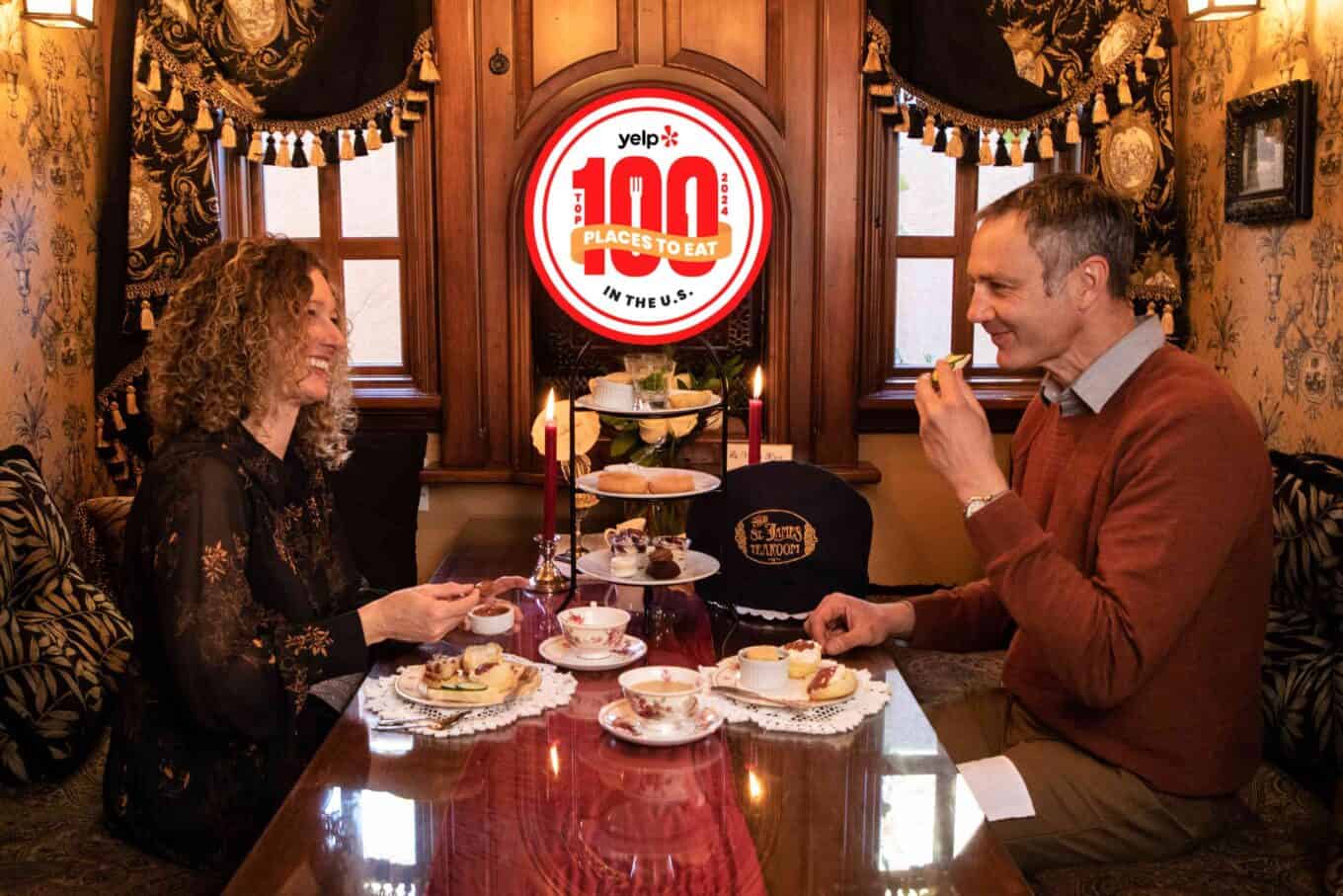 A man and woman enjoy tea and pastries at a decorated table in a cozy room, with a “Yelp’s Top 100 Restaurants” sign proudly displayed in the window behind them.