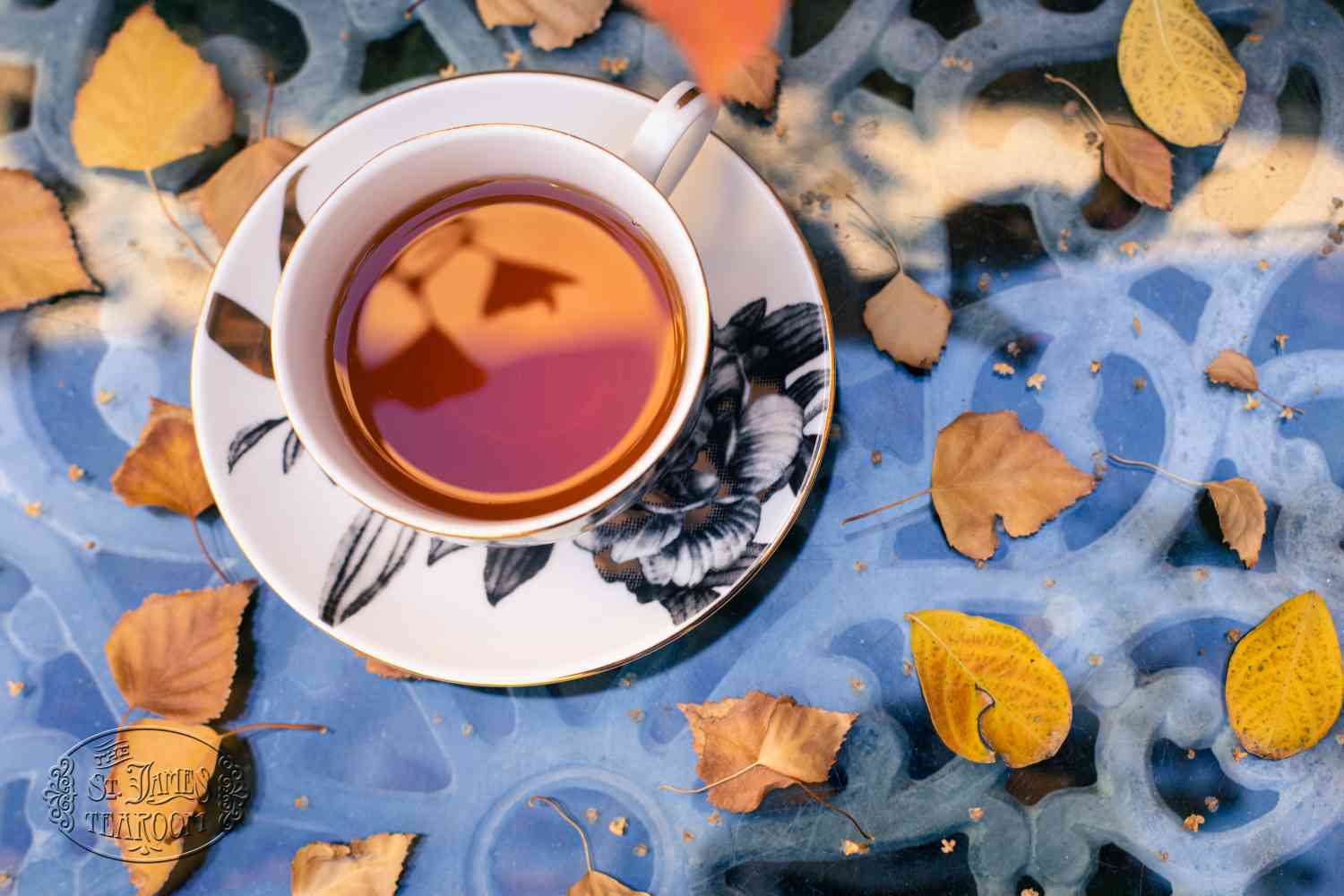 A cup of tea sits on a saucer with a floral design, placed on a blue glass table scattered with yellow and brown autumn leaves, inviting you to savor the harvest in every soothing sip.