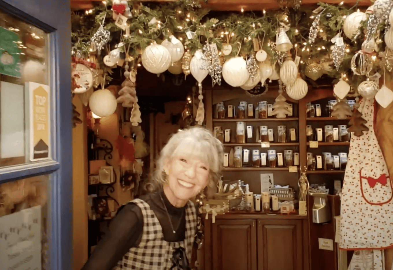 A smiling woman stands in a cozy, festive shop decorated with ornaments, twinkling lights, and holiday greenery. Shelves in the background hold various small products. A red and white apron hangs nearby.