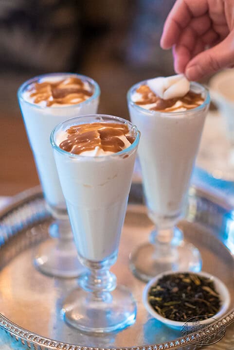 Three glasses of creamy dessert with caramel drizzles on top are placed on a silver tray. A hand is adding a topping to one glass. A small dish with loose tea leaves sits nearby.