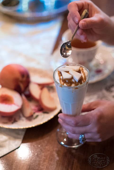 A hand holds a glass of yogurt topped with caramel and coconut, while another hand spoons some topping. Sliced peaches rest on a plate beside a teacup on a wooden table.