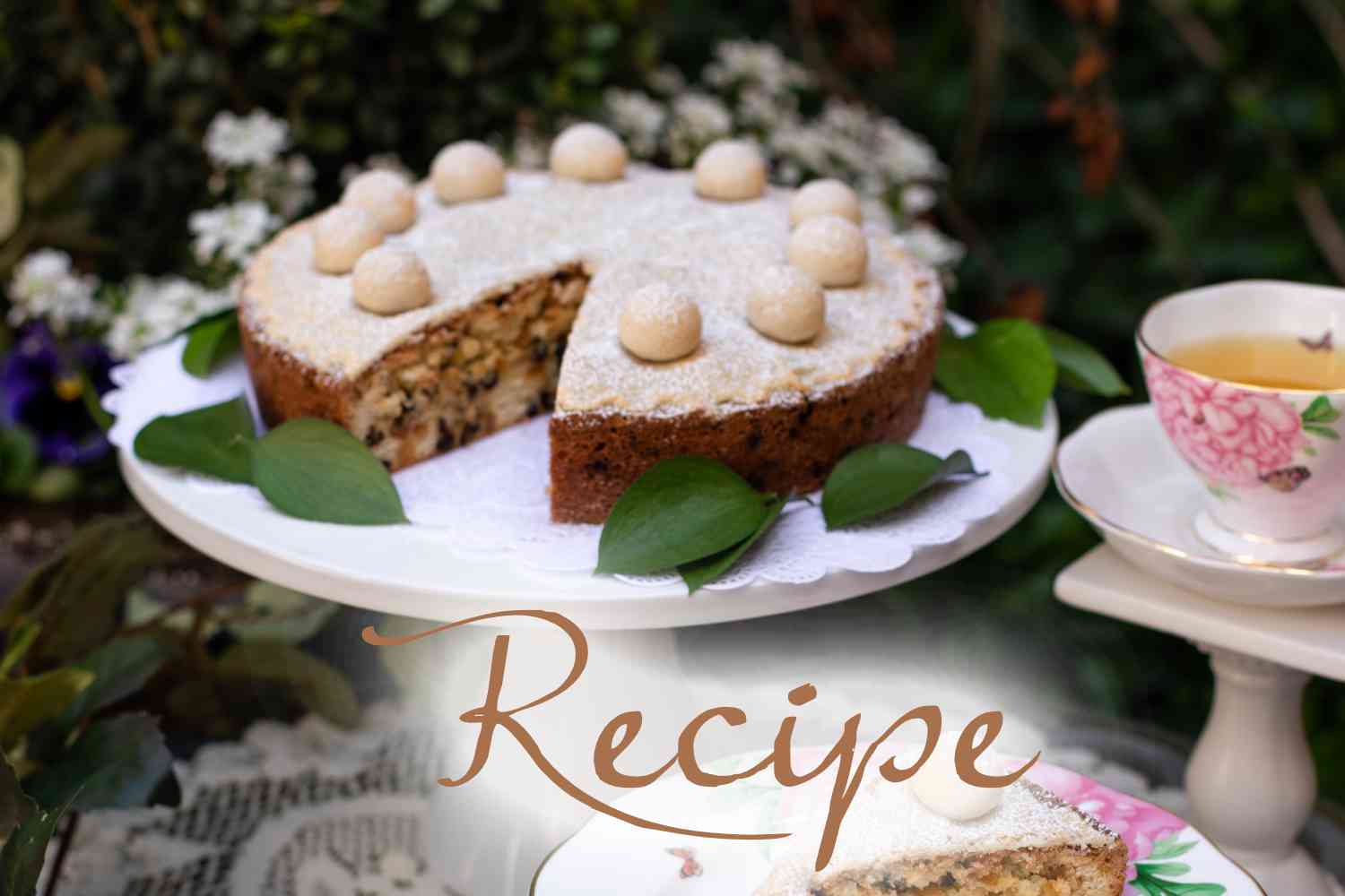 A round Simnel Cake, perfect for a special Simnel Cake Recipe for Mother’s Day, sits with a slice missing, topped with small marzipan balls and powdered sugar on a lace doily adorned with green leaves. A teacup and saucer rest beside it. “Recipe” is written across the image.
