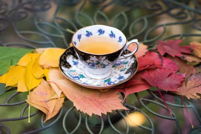 A floral teacup filled with tea sits on an ornate glass table, surrounded by colorful autumn leaves in shades of yellow, orange, and red.