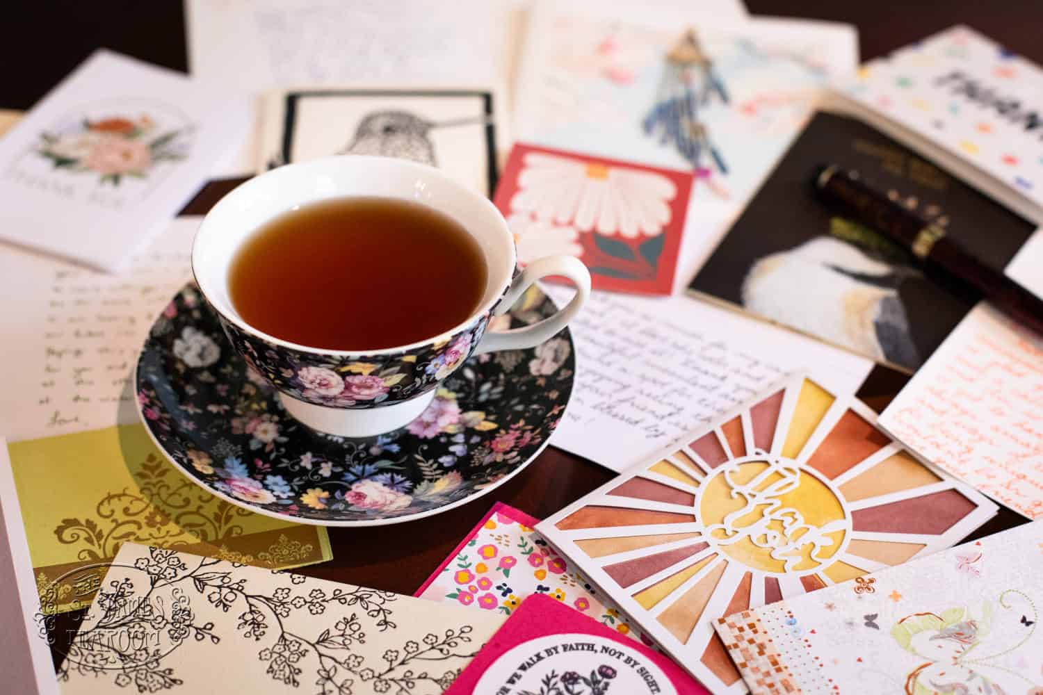 A floral teacup filled with tea sits on a matching saucer, surrounded by handwritten letters, cards, and colorful envelopes on a table—a charming scene that reflects the timeless art of caring for fine china.