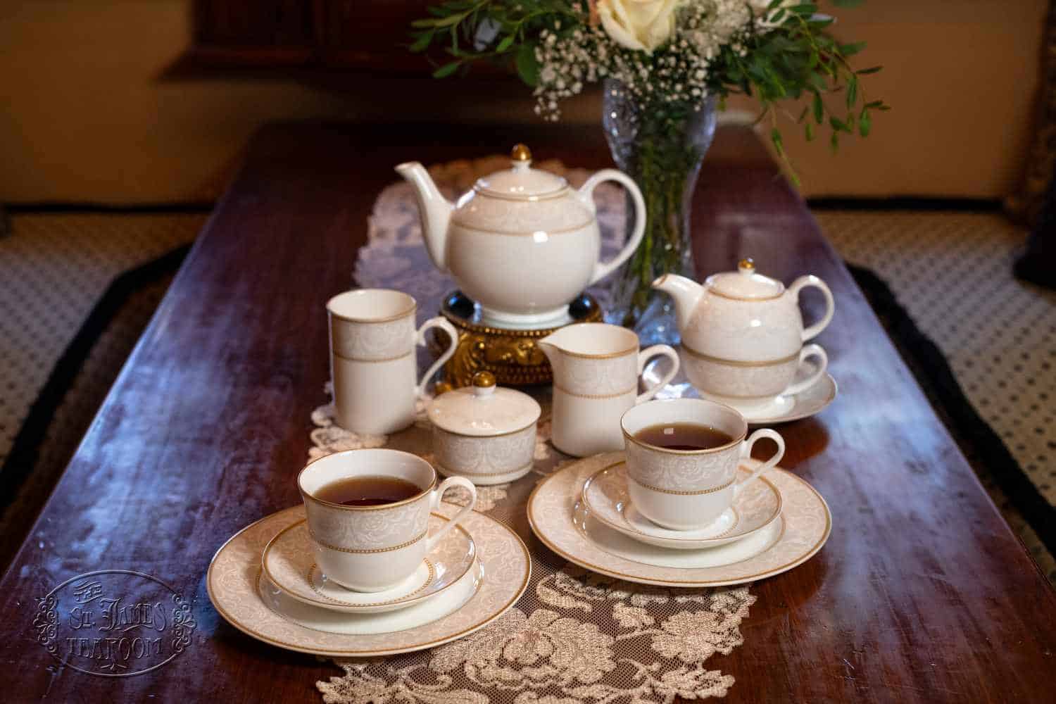 Elegant tea set with white and gold cups, saucers, teapots, and a vase of flowers, arranged neatly on a wooden table with a lace runner. Two cups are filled with tea, reflecting the thoughtful art of caring for fine china in every detail of this beautifully presented scene.