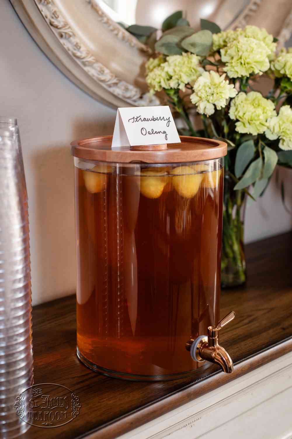 A glass beverage dispenser filled with strawberry oolong iced tea and lemon slices, inspired by a refreshing Strawberry Iced Tea Recipe, sits on a wooden table next to a stack of clear cups and a vase of light green flowers.
