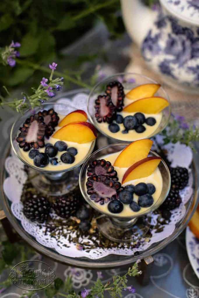 Three glass cups filled with creamy dessert, inspired by a Tea Infused Creme Anglaise recipe, are topped with blackberries, blueberries, and peach slices. They sit on a decorative tray surrounded by flowers and a teapot.