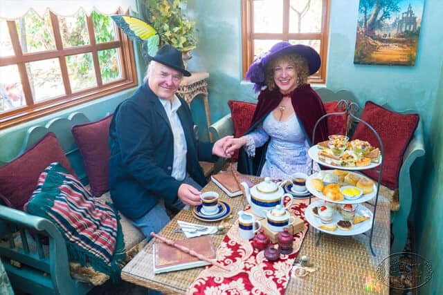 A smiling couple dressed in vintage attire enjoys afternoon tea at a cozy, ornate table with a tiered tray of treats, teapots, and cups. They hold hands and sit by a window in a warmly decorated room.