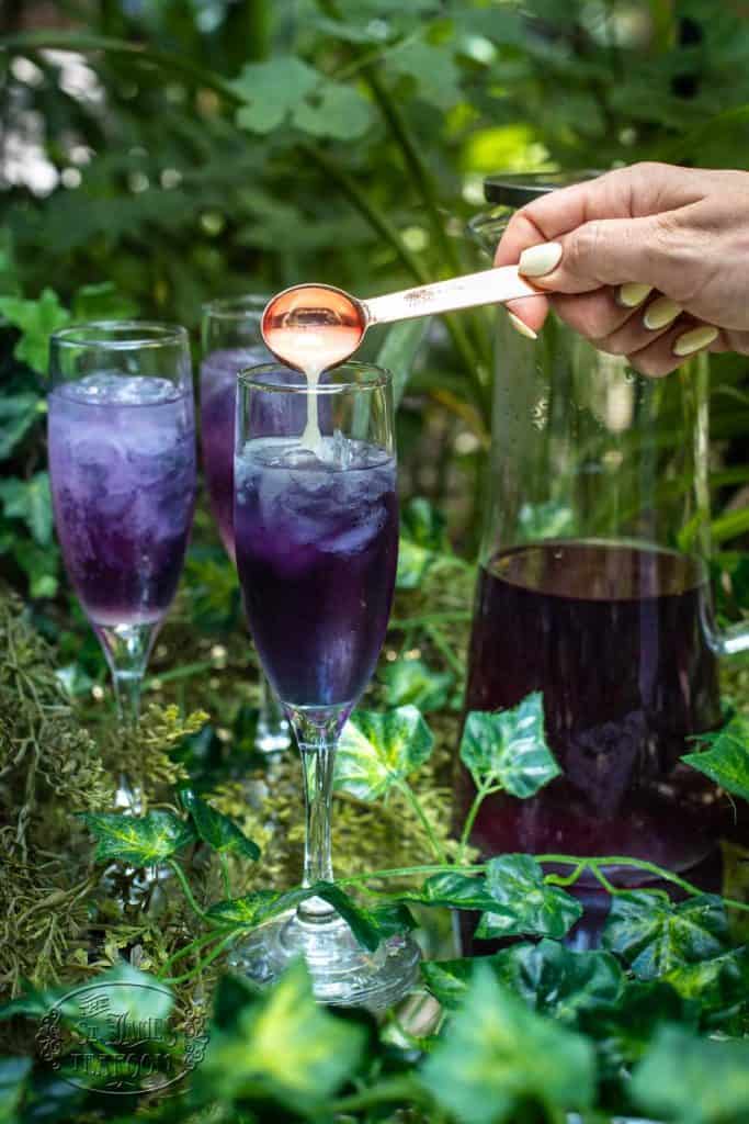 A hand pours syrup from a spoon into a tall glass of purple iced drink, surrounded by greenery, with more glasses and a pitcher in the background, creating an inviting scene that hints at a refreshing Iced Tea Recipe perfect for summer gatherings.