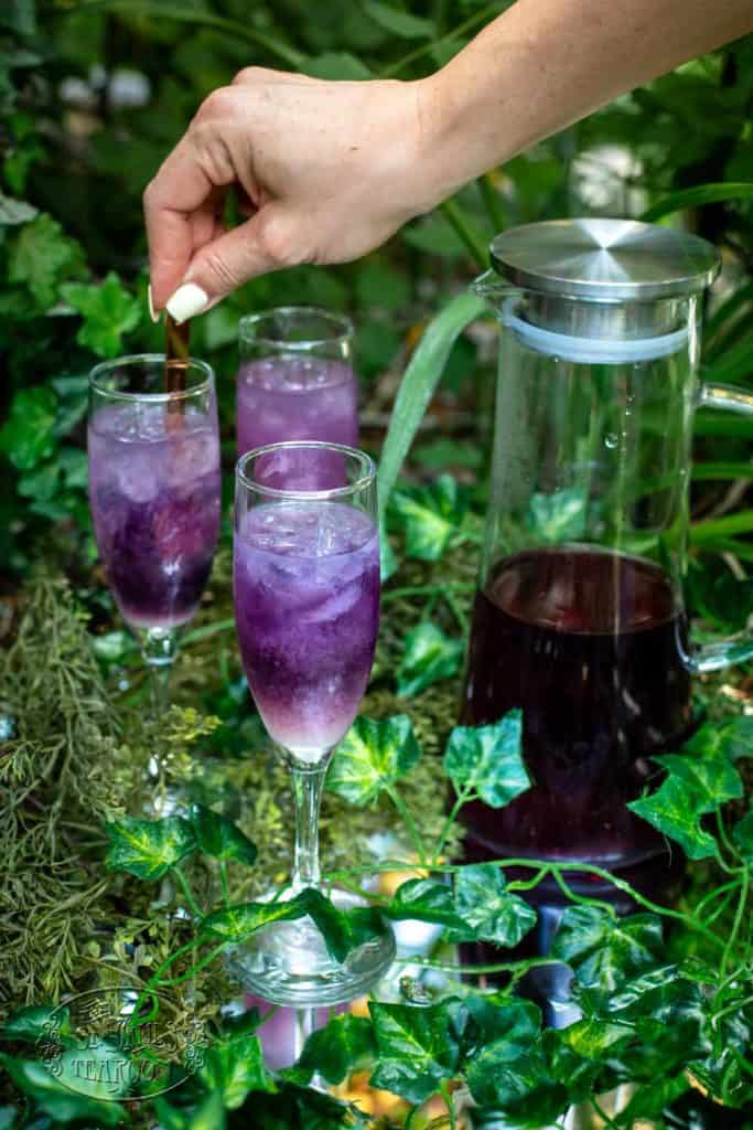 A hand stirs a vibrant purple iced drink in a champagne glass, showcasing an elegant twist on an Iced Tea Recipe. Two more glasses and a pitcher of the same refreshing beverage sit on a table surrounded by green ivy and foliage.