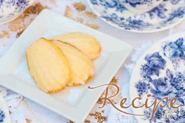 Three golden madeleines rest on a white square plate, surrounded by teacups and saucers adorned with blue floral patterns. The word “Recipe” is written in script across the image, perfectly capturing the charm of homemade madeleines.