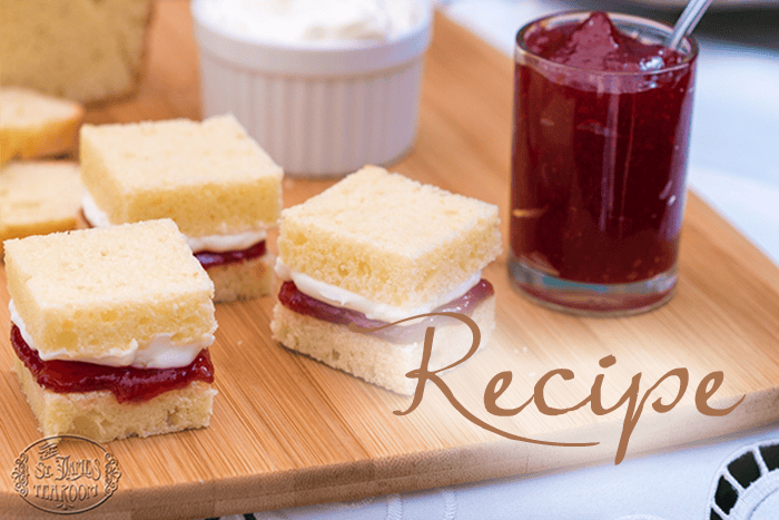 Small square Victoria Sponge Cakes with cream and red jam filling rest on a wooden board, accompanied by a glass of jam and a bowl of cream nearby. The word “Recipe” is overlaid on the image.