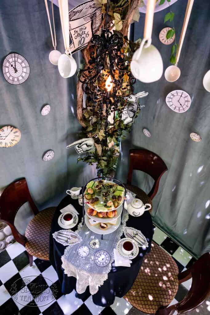 A whimsical tea party setup with mismatched clocks, teacups hanging above, a tree centerpiece, black-and-white checkered floor, and a table set with tea, pastries, and lace. Alice in Wonderland-themed decor surrounds the scene.