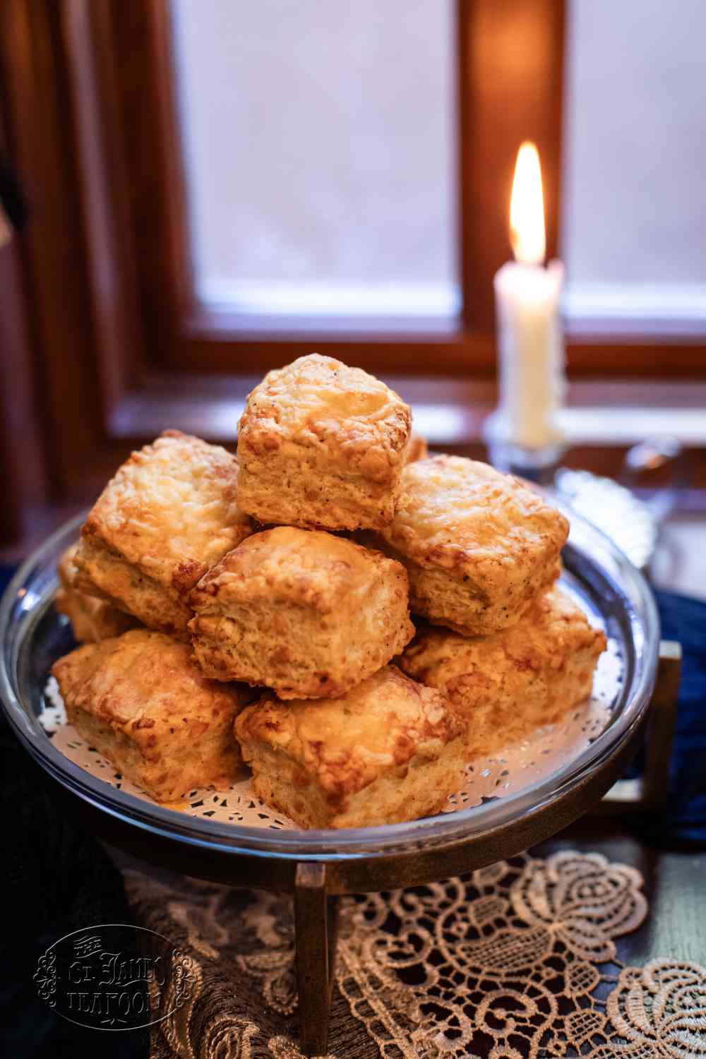 A plate of flaky, golden biscuits—reminiscent of a classic White Cheddar Black Pepper Scones recipe—is stacked in a pyramid atop a lace doily, with a lit candle and a window in the softly blurred background.