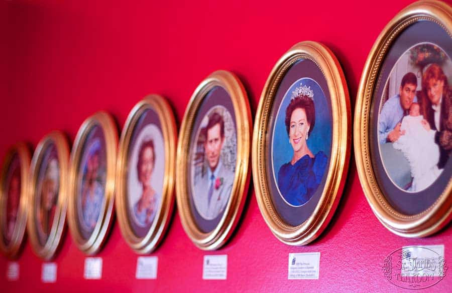 A row of oval-framed portraits hangs on a bright red wall, showing various people, including men and women, in both formal and casual attire. The frames are gold, and each portrait has a small label beneath it.