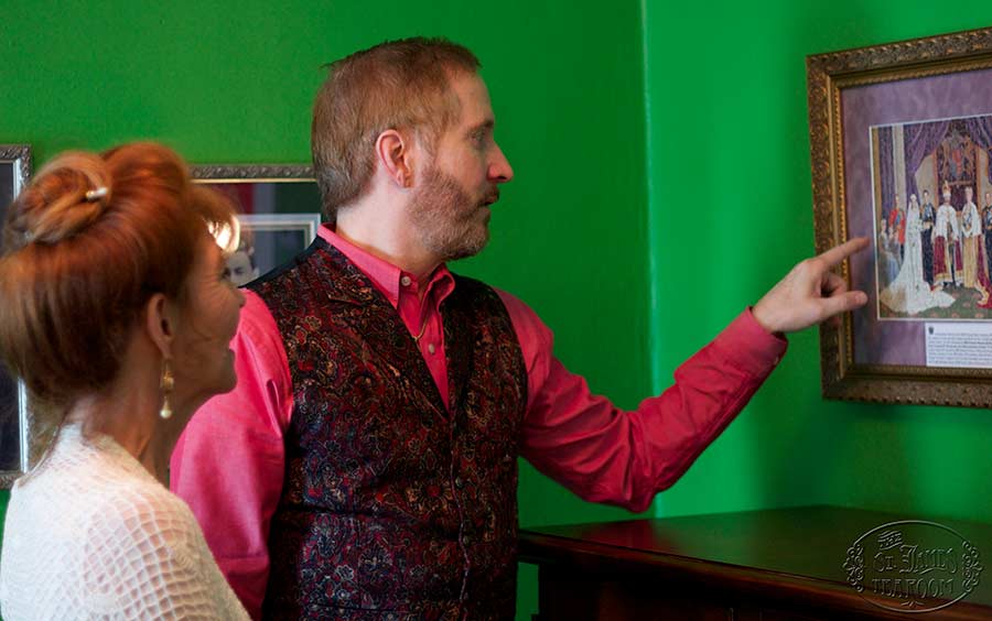 A man in a red shirt and patterned vest points at a framed artwork on a green wall while a woman with red hair stands beside him, both looking at the picture.