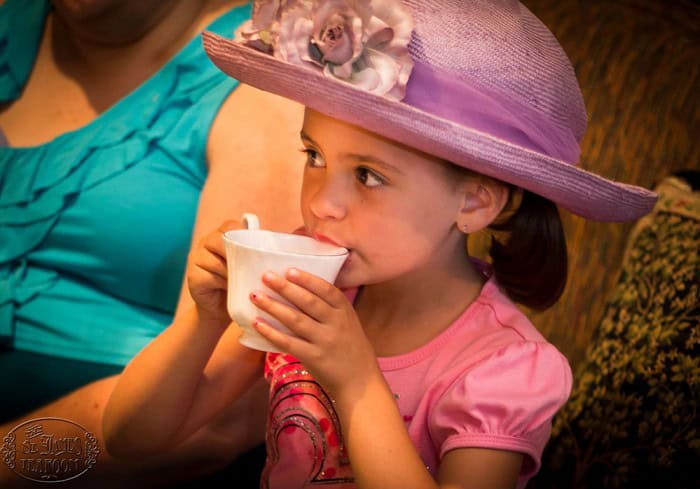 A young girl wearing a lavender wide-brimmed hat with a flower drinks from a white teacup. She is dressed in a pink shirt and is sitting next to an adult in a teal top.