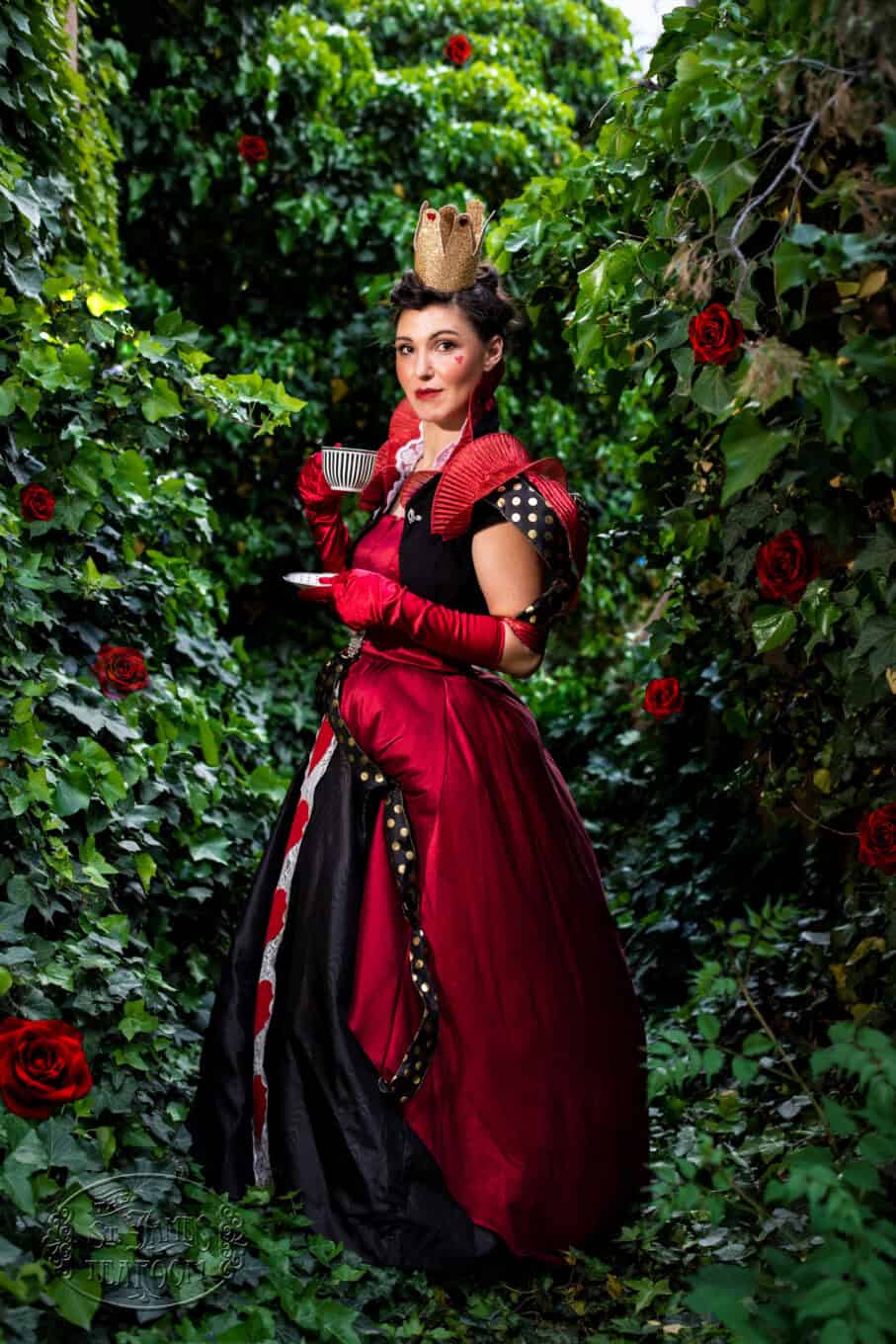 A woman in a red and black queen costume with a crown holds a teacup and saucer while standing in a lush, leafy garden with red roses.
