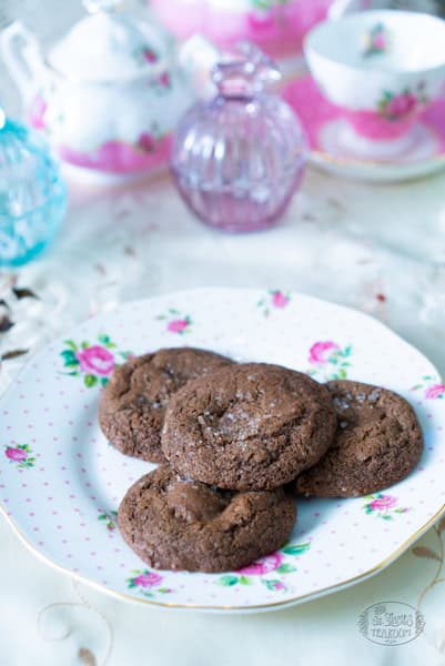 Four chocolate cookies are arranged on a floral-patterned plate. In the background, pastel-colored glassware and china teacups add a delicate, vintage tea party atmosphere, perfectly complementing the irresistible charm of the chocolate cookies.
