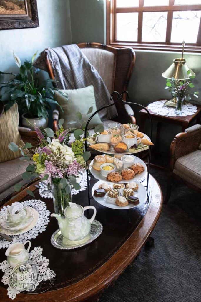 A cozy tea room with armchairs, a wooden table set with a teapot, teacups, a vase of flowers, and a three-tiered tray of pastries and treats. A lamp and lace doilies add a vintage touch.
