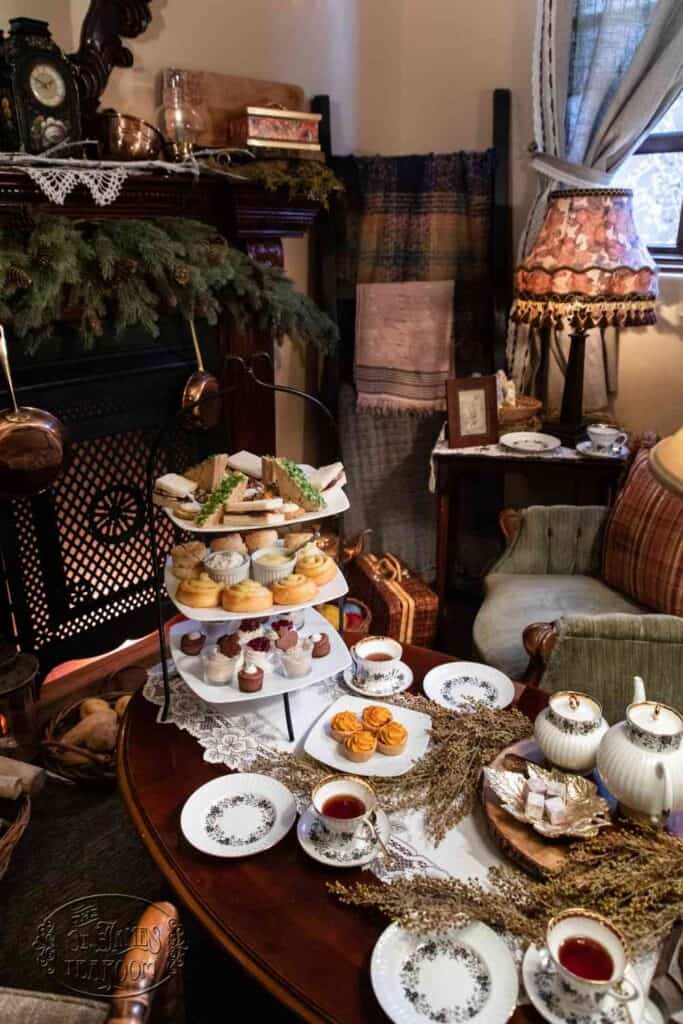 A cozy tea setting with a tiered tray of pastries, teapots, teacups, and plates on a wooden table, surrounded by vintage furniture, blankets, and warm decorations in a softly lit room.