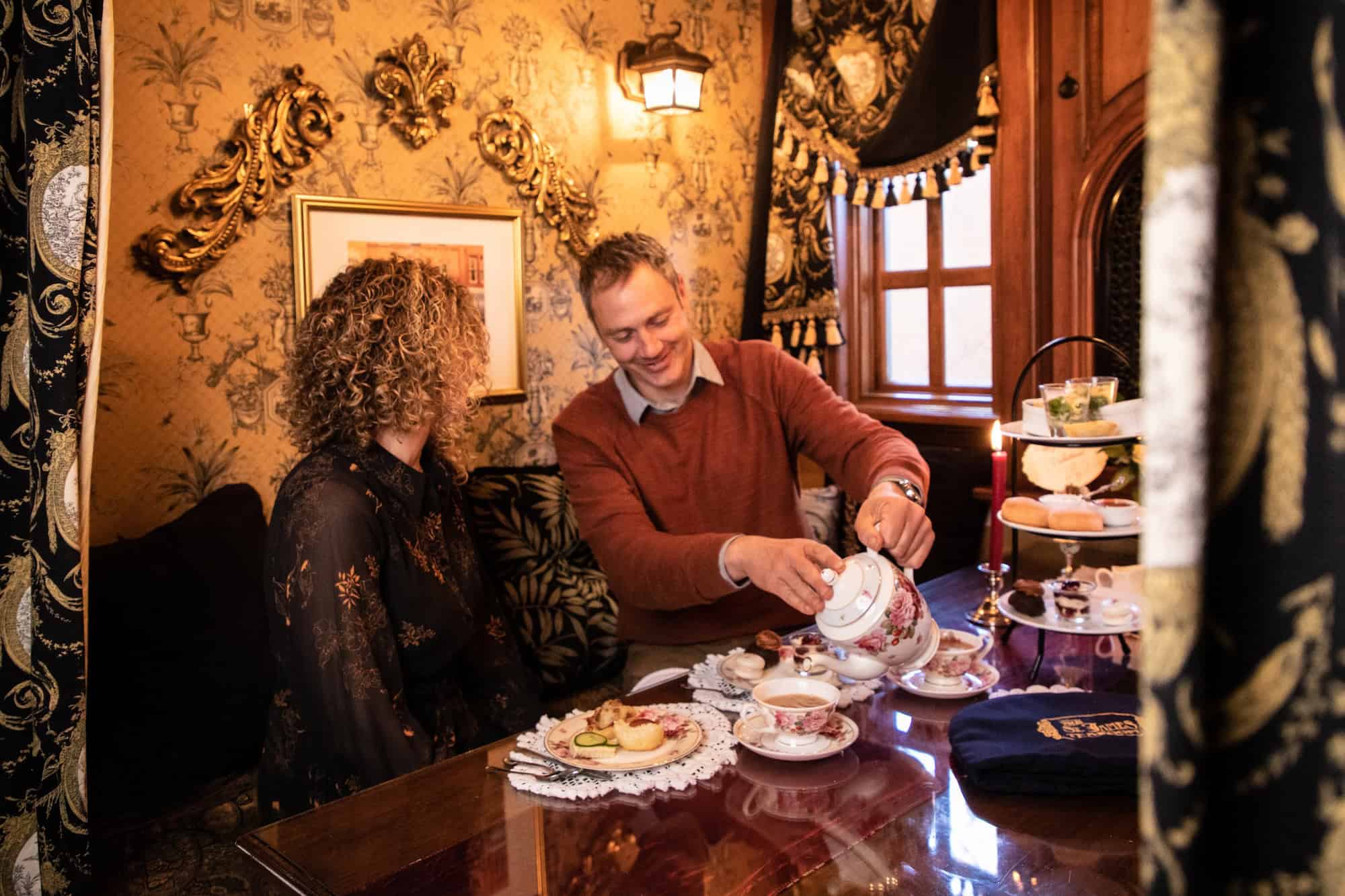 A man pours tea for a woman at an ornate, cozy table set with pastries and treats, surrounded by gold-patterned wallpaper and elegant decor.