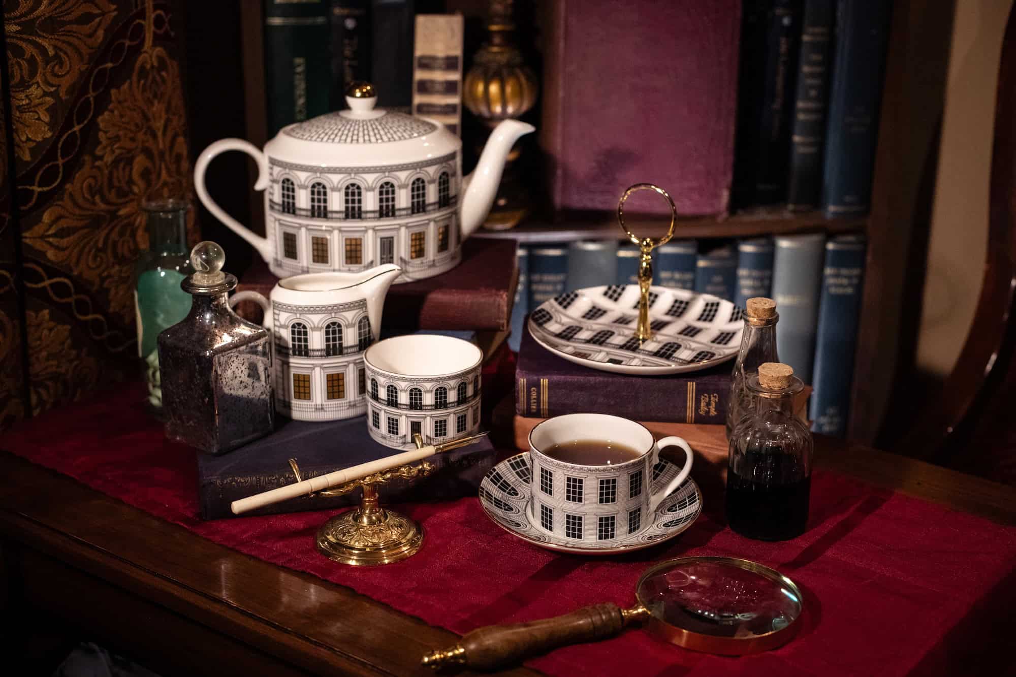 A tea set with architectural designs, books, glass bottles, a magnifying glass, a pen, and a sealed ink bottle are arranged on a red cloth, with shelves of books in the background.