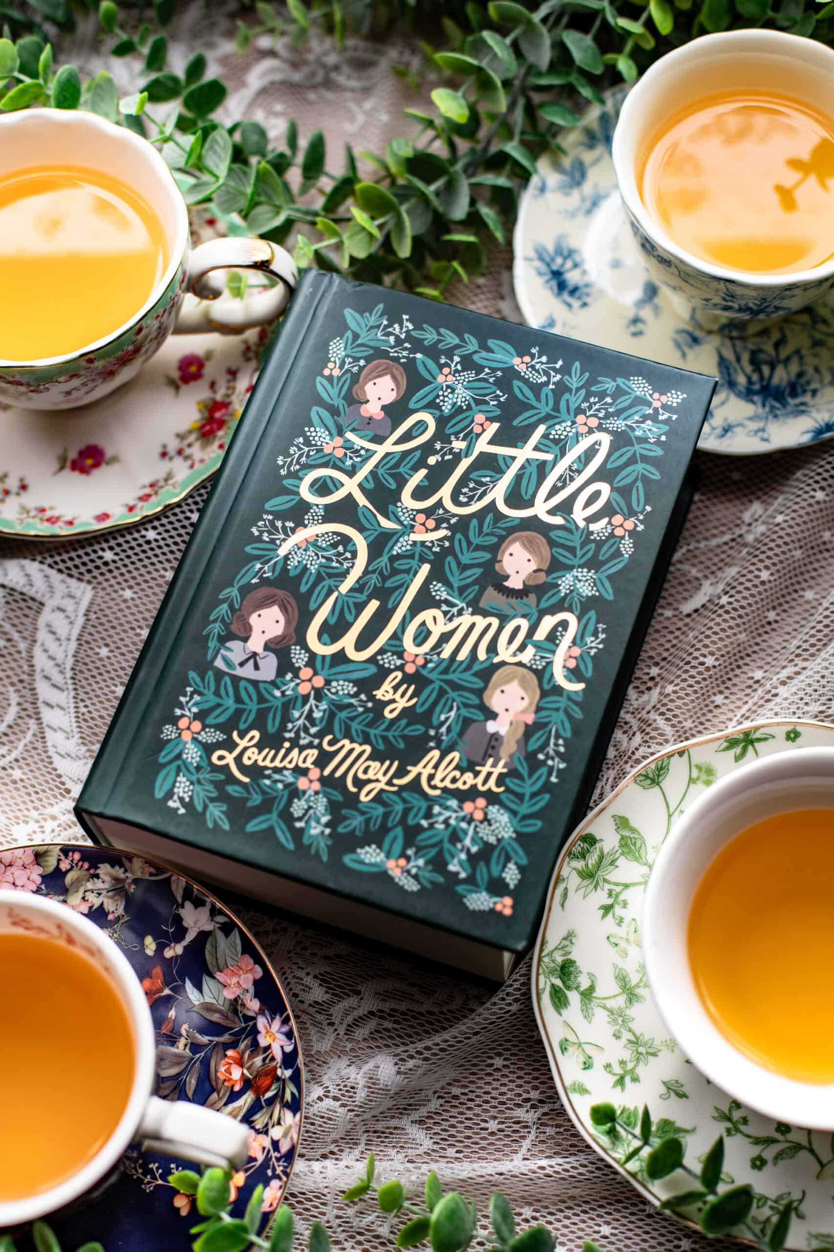 A copy of “Little Women” by Louisa May Alcott lies on lace fabric, surrounded by teacups filled with tea and leafy greenery.