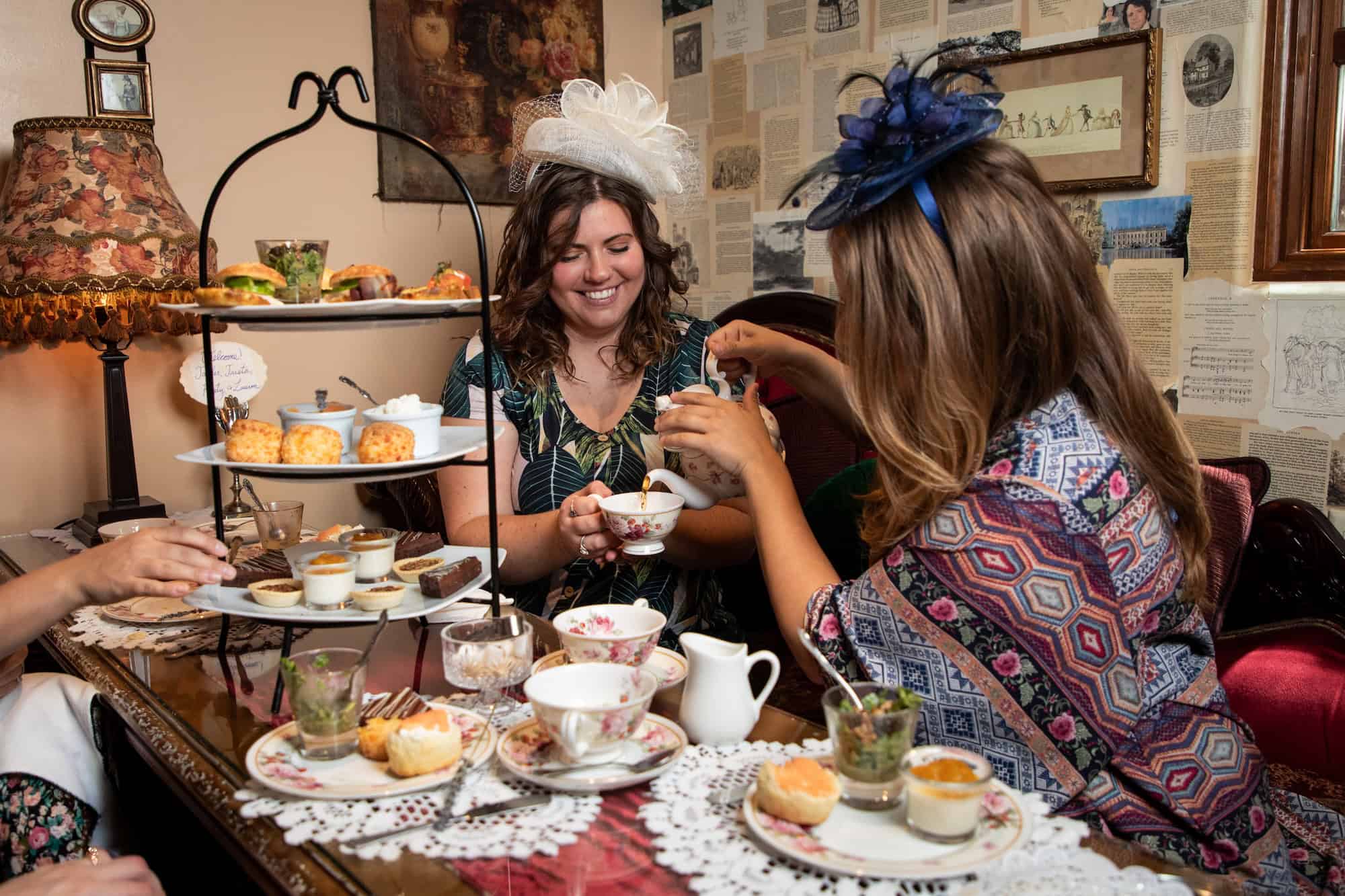 Two women wearing fancy hats enjoy afternoon tea with pastries, scones, and desserts on a tiered tray in a cozy, vintage-themed room decorated with old book pages and framed pictures.