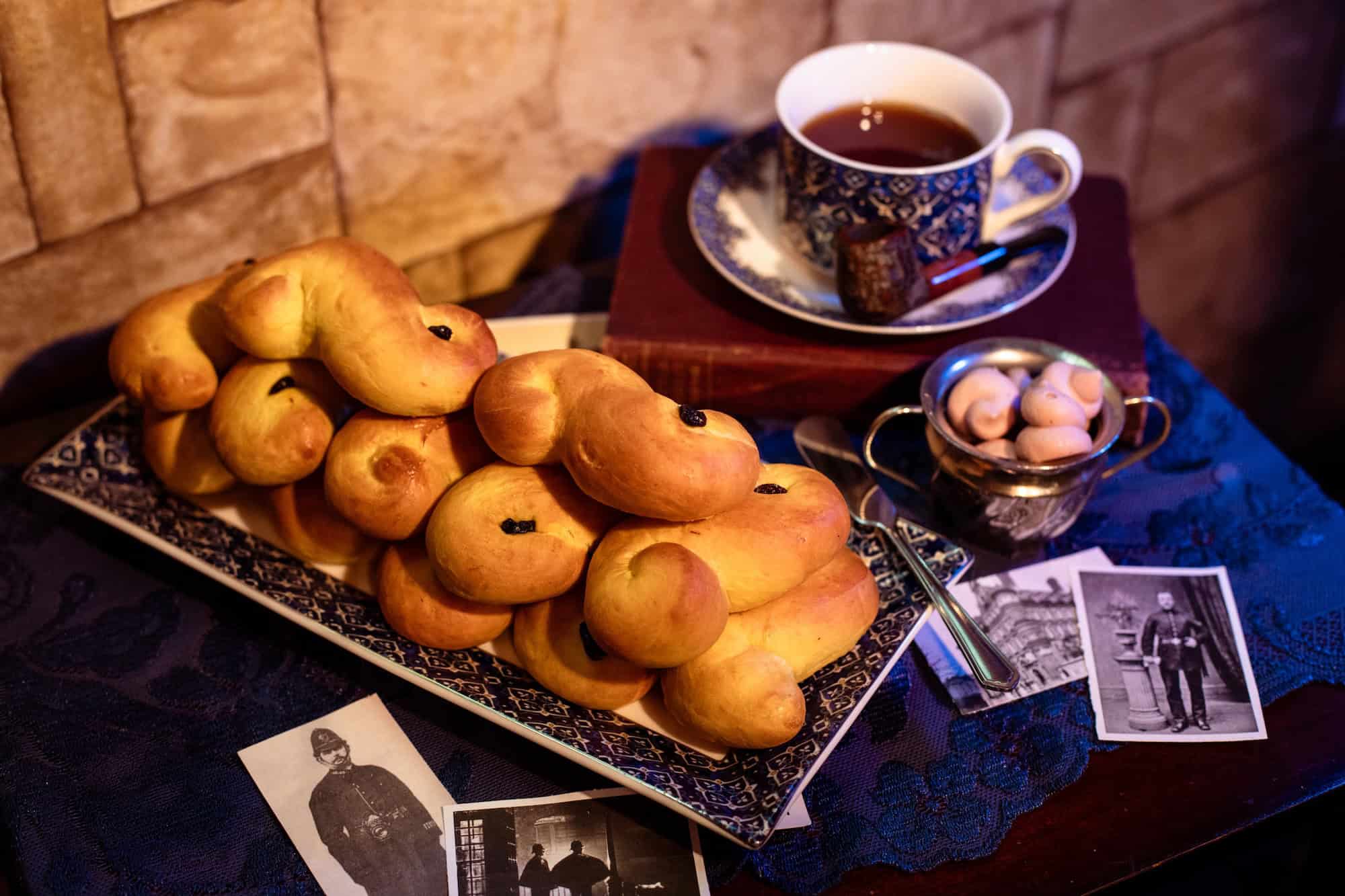A plate of saffron buns, a teacup on a book, a small bowl of cookies, and vintage black-and-white photos on a table with a blue cloth and a tiled wall background.