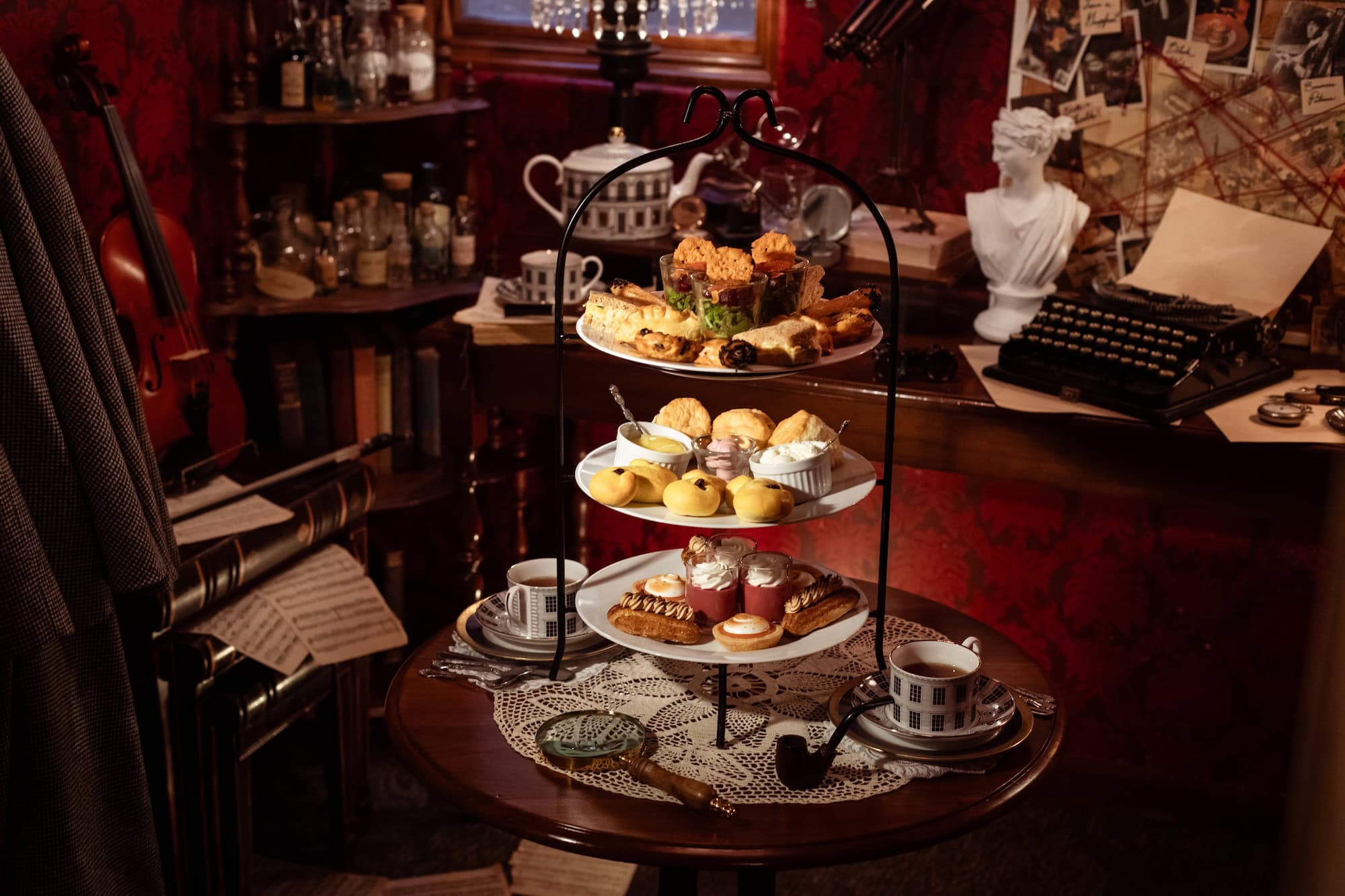 A three-tiered tray with assorted pastries and sandwiches sits on a round table with lace, teacups, and a magnifying glass in a vintage, cluttered study with a typewriter, bust, and violin in the background.