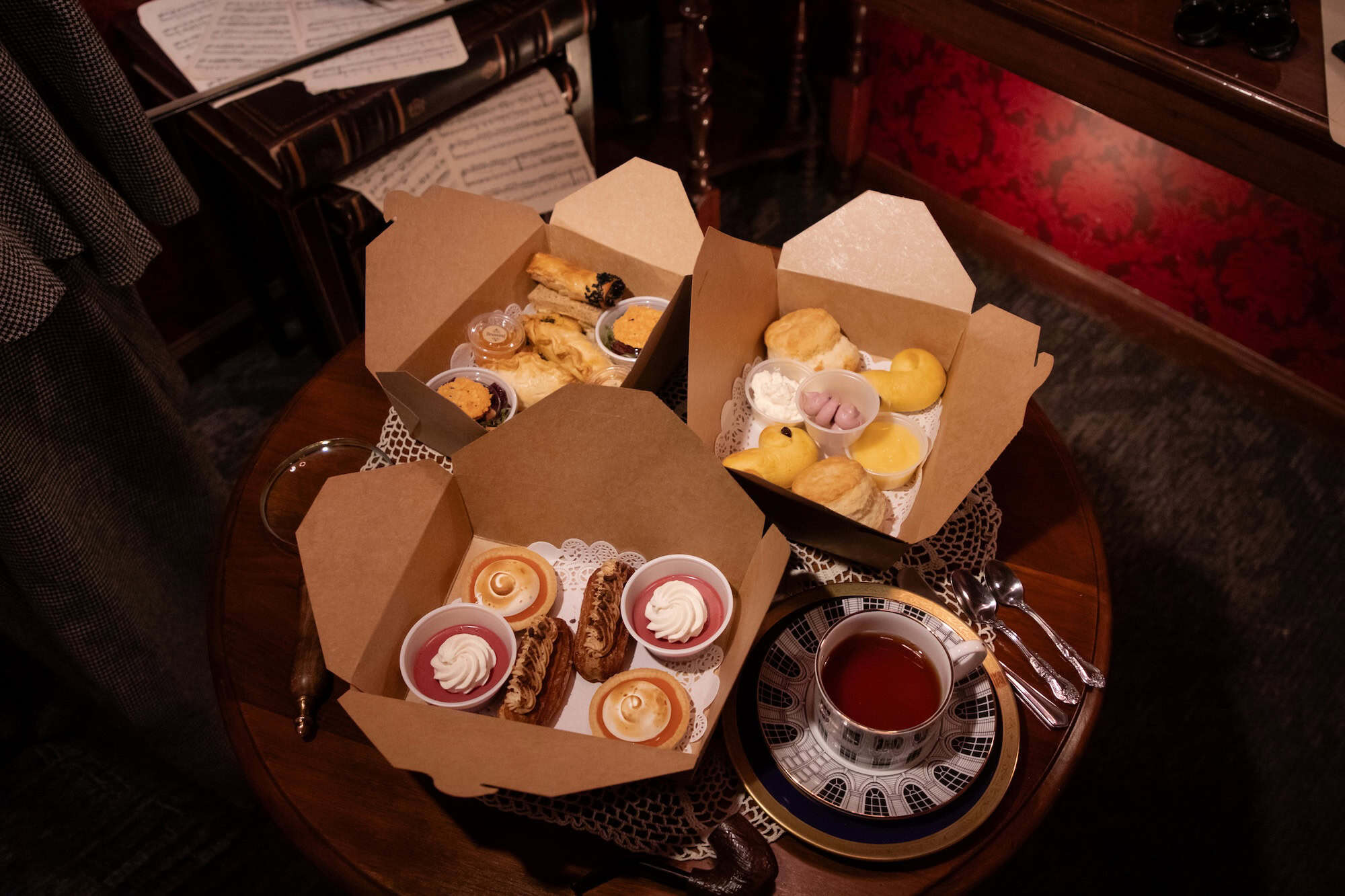 Three open takeout boxes with assorted pastries, cream, and scones sit on a round wooden table next to a cup of tea, silverware, and sheet music in a cozy, warmly lit setting.