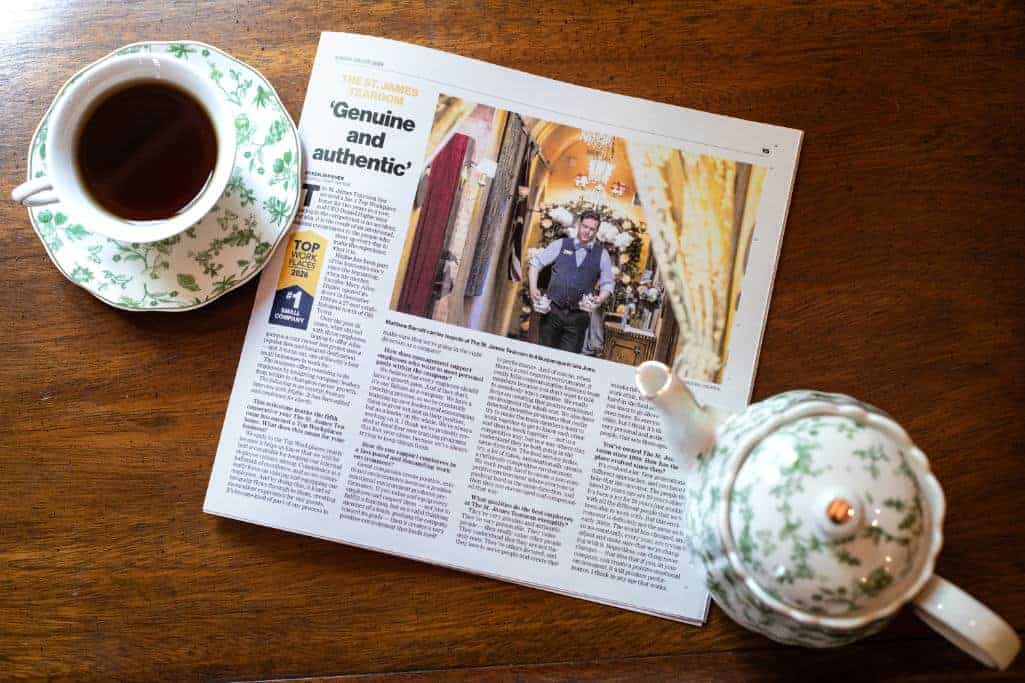 A newspaper article on a wooden table beside a cup of tea and a floral-patterned teapot and saucer. The article features a photo of a man standing in a warmly lit room.