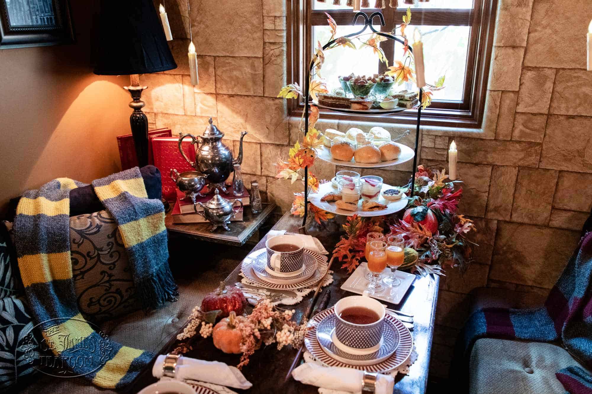 A tea table set for two with a three-tiered tray of sandwiches, scones, and desserts, decorated with autumn leaves and pumpkins.