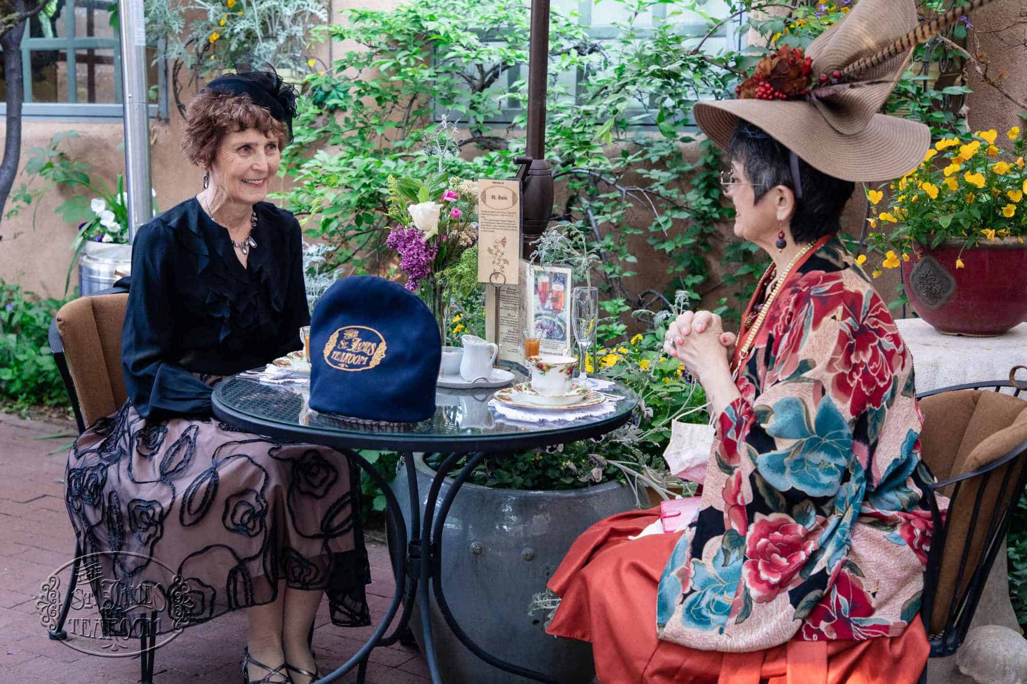 Two women wearing hats sit at an outdoor table for tea in a garden setting.