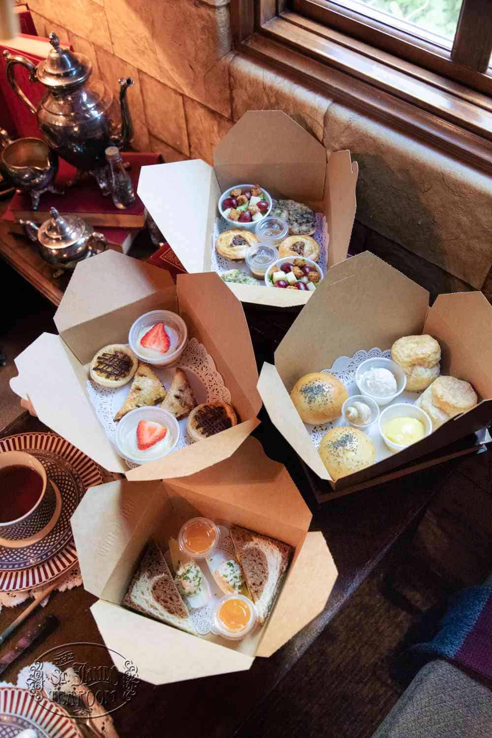 Four open takeout boxes containing tea party food, including sandwiches, scones with cream, fruit, and pastries, arranged on a table with a teacup.
