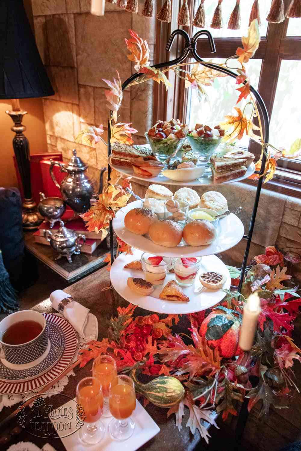A three-tiered tea service featuring finger sandwiches, scones, and small desserts, decorated with autumn leaves and surrounding tea service items.