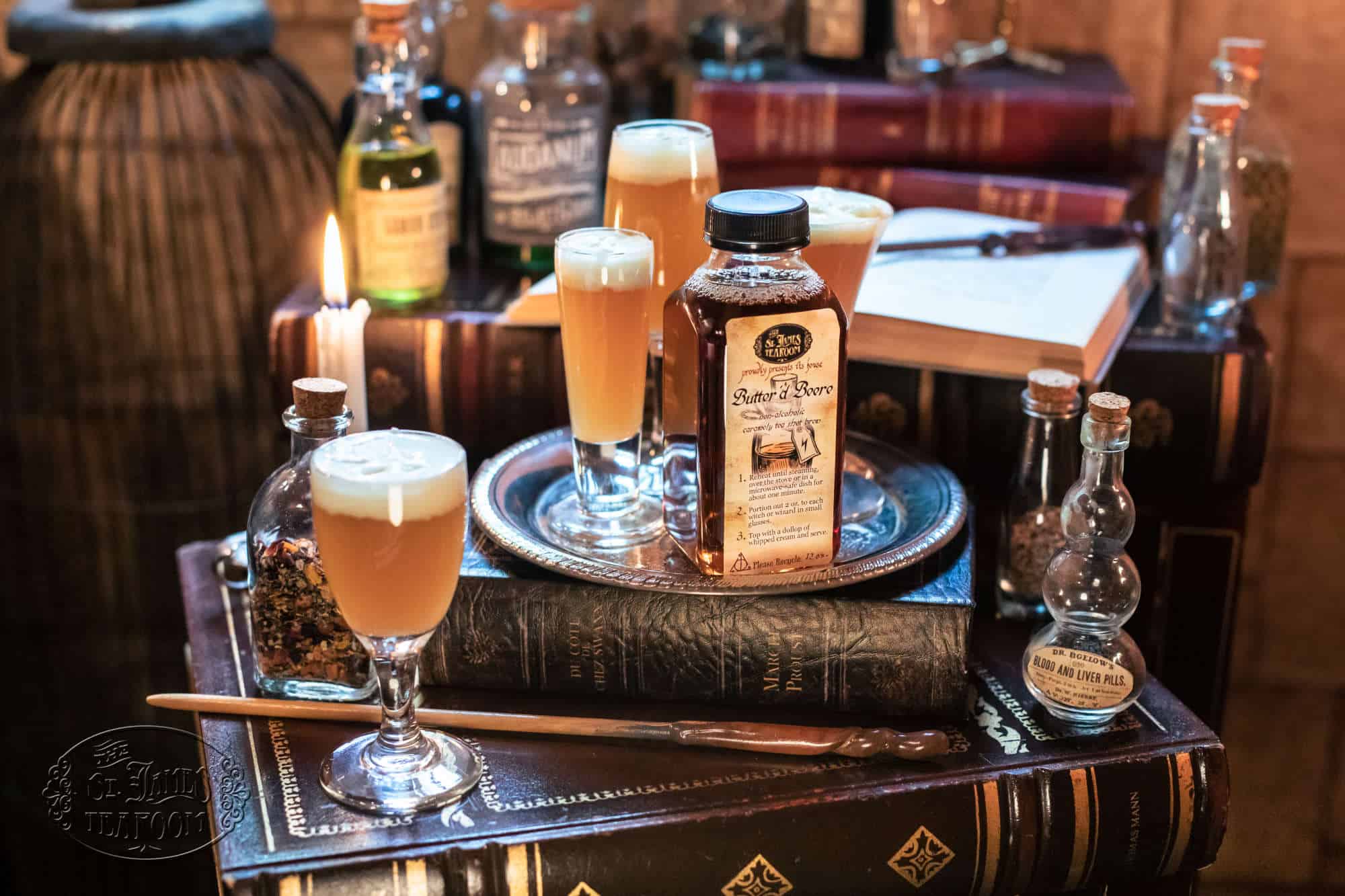 A bottle of Butter'd Beere labeled with brewing instructions, surrounded by filled glasses, potion bottles, an open book, and a wand on a stack of old books.